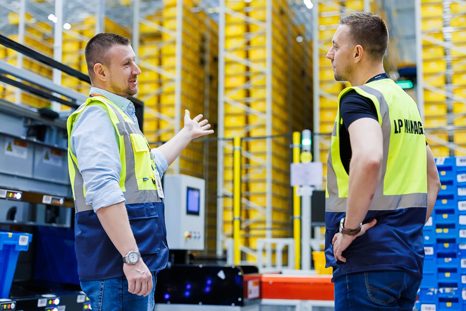 Dwaj pracownicy magazynu w kamizelkach bezpieczeństwa omawiają procesy przy regałach z żółtymi pojemnikami
