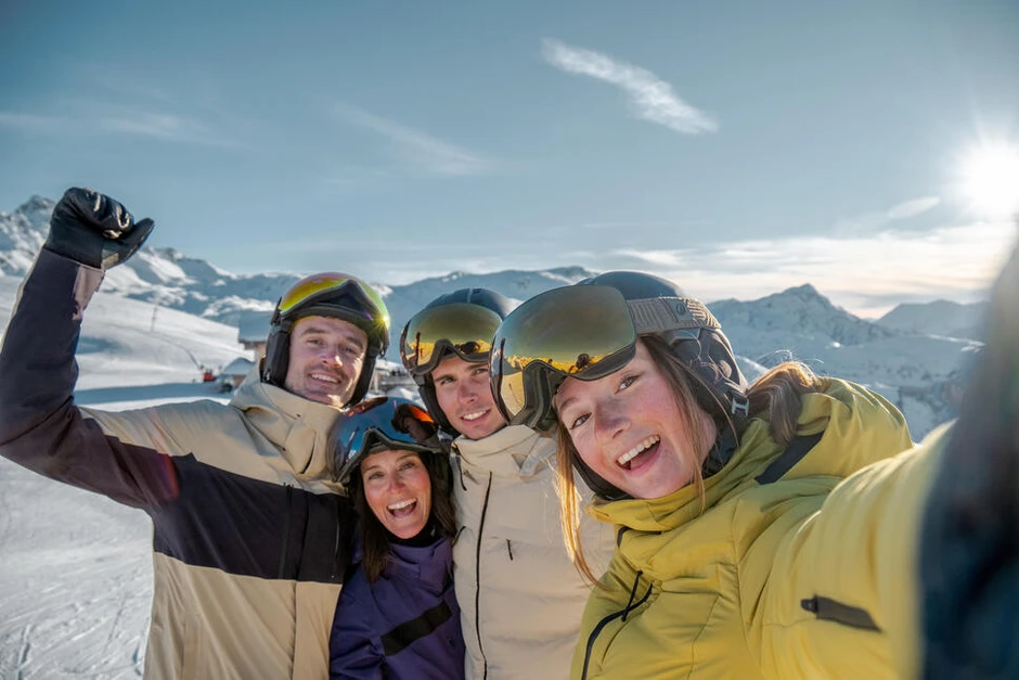 Grupa uśmiechniętych osób w strojach narciarskich na śniegu robiąca wspólne selfie na tle gór