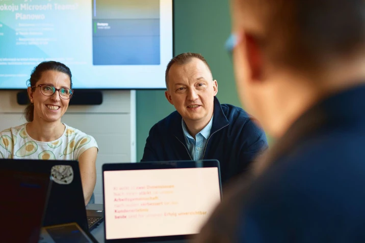 Dwóch pracowników rozmawia podczas spotkania w nowoczesnej sali konferencyjnej z widocznym ekranem i laptopami