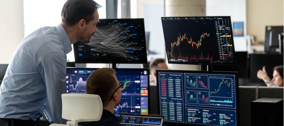 Dwóch mężczyzn analizuje dane i wykresy na wielu monitorach w nowoczesnym biurze firmy finansowej