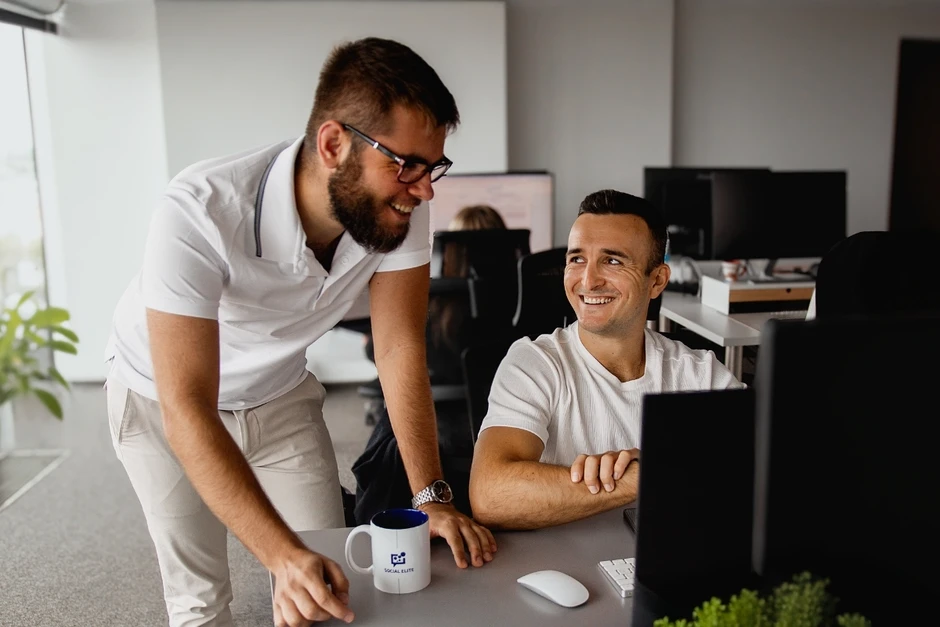 Dwóch uśmiechniętych mężczyzn współpracuje przy biurku w nowoczesnym biurze z komputerami