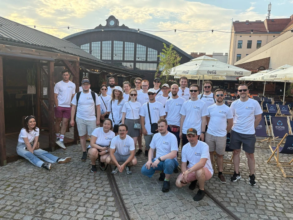 Grupa młodych ludzi w białych koszulkach Apollo Poland 2025 pozujący na zewnątrz przy brukowanym placu
