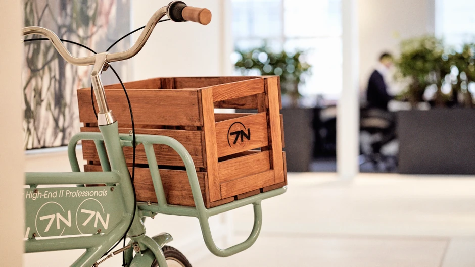 Rower z drewnianą skrzynią i logo firmy w nowoczesnym biurze z roślinami i pracownikiem w tle