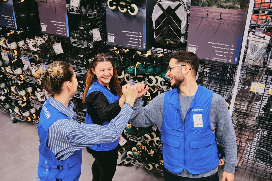 Trójka pracowników Decathlon w niebieskich kamizelkach wymienia się uśmiechami i uściśnięciami dłoni w sklepie z rolkami