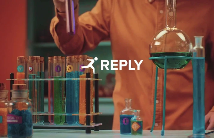 Laboratoryjne probówki z kolorowymi cieczami w tle osoby w pomarańczowej koszuli z logo Reply