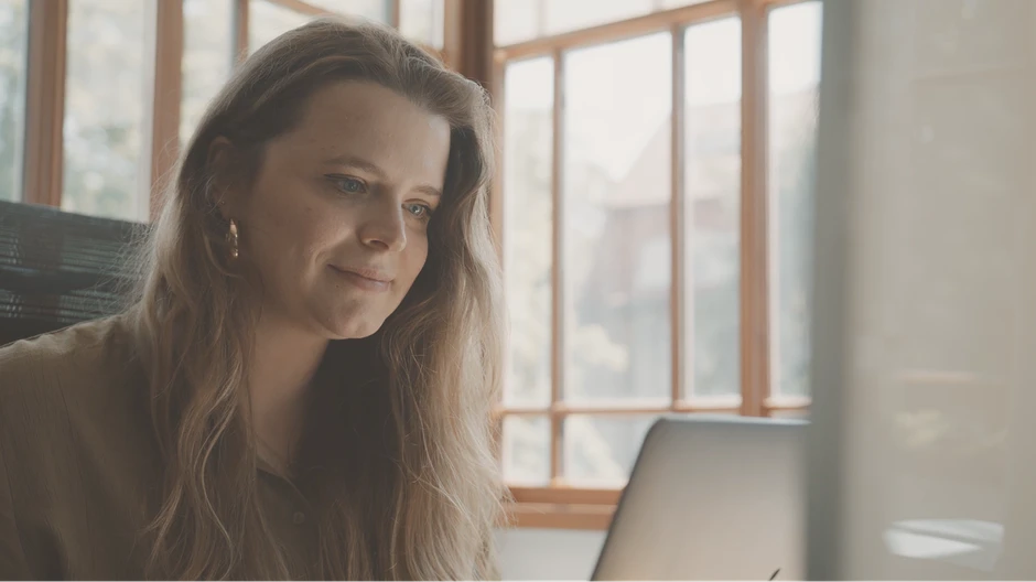 Młoda kobieta pracuje przy laptopie w jasnym biurze z dużym oknem i drewnianą ramą