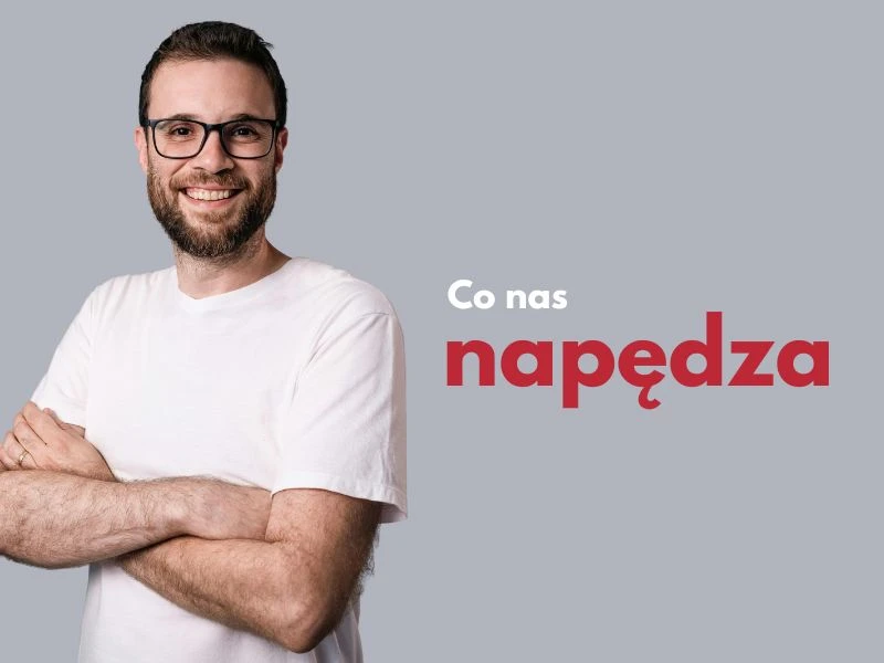 Uśmiechnięty mężczyzna w białej koszulce z założonymi rękami na szarym tle z napisem Co nas napędza
