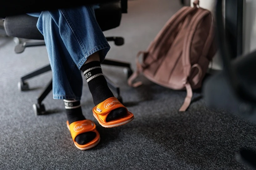 Osoba siedząca na krześle biurowym w dżinsach i pomarańczowych klapkach na czarnych skarpetach na wykładzinie biurowej