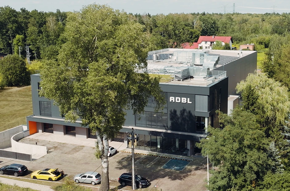 Nowoczesny budynek firmy ADBL otoczony zielenią i miejscami parkingowymi przy słonecznej pogodzie