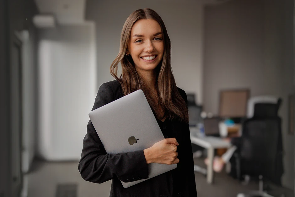 Młoda kobieta w czarnej marynarce trzyma laptopa i uśmiecha się stojąc w nowoczesnym biurze