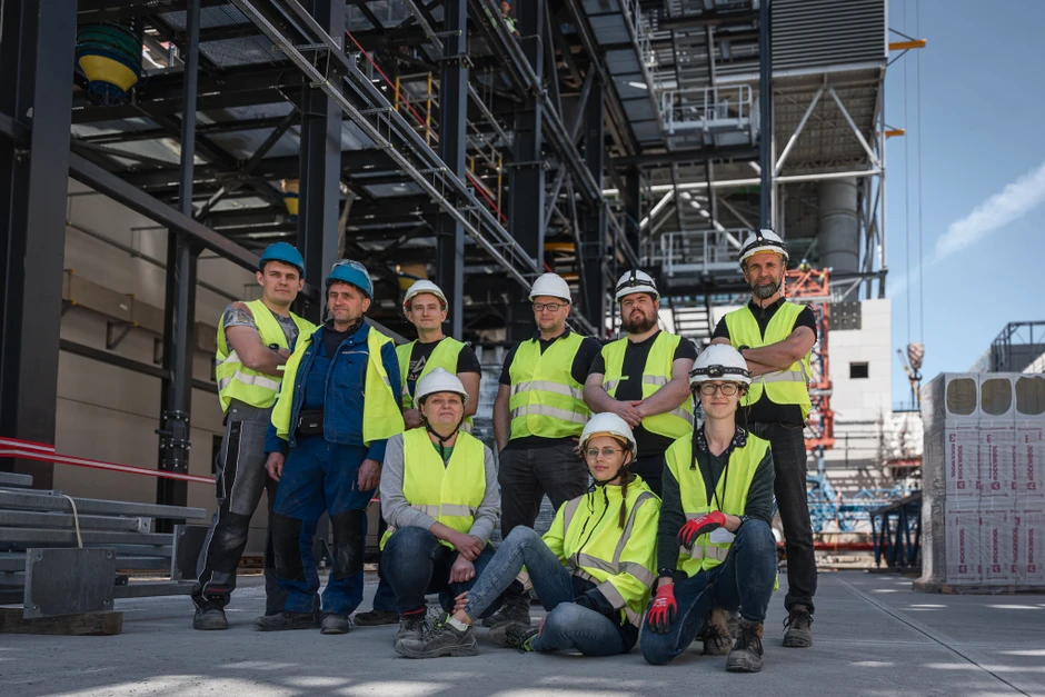 Grupa pracowników budowlanych w kamizelkach odblaskowych i kaskach stoi na terenie placu budowy przy nowoczesnym obiekcie