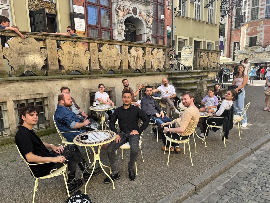 Grupa osób spotyka się na zewnątrz przy stolikach kawiarni w historycznym centrum miasta