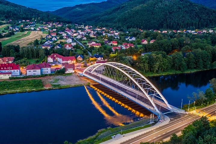 Nowoczesny most łukowy nad rzeką w malowniczej miejscowości otoczonej zielonymi wzgórzami i domami