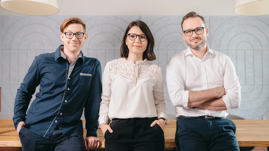 Trójka młodych profesjonalistów w biurze pozująca do zdjęcia na tle jasnej ściany z dekoracją