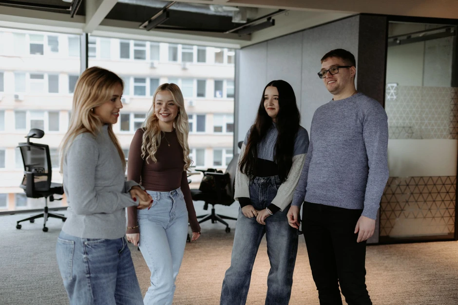 Grupa młodych pracowników rozmawia i uśmiecha się w nowoczesnym biurze z dużymi oknami