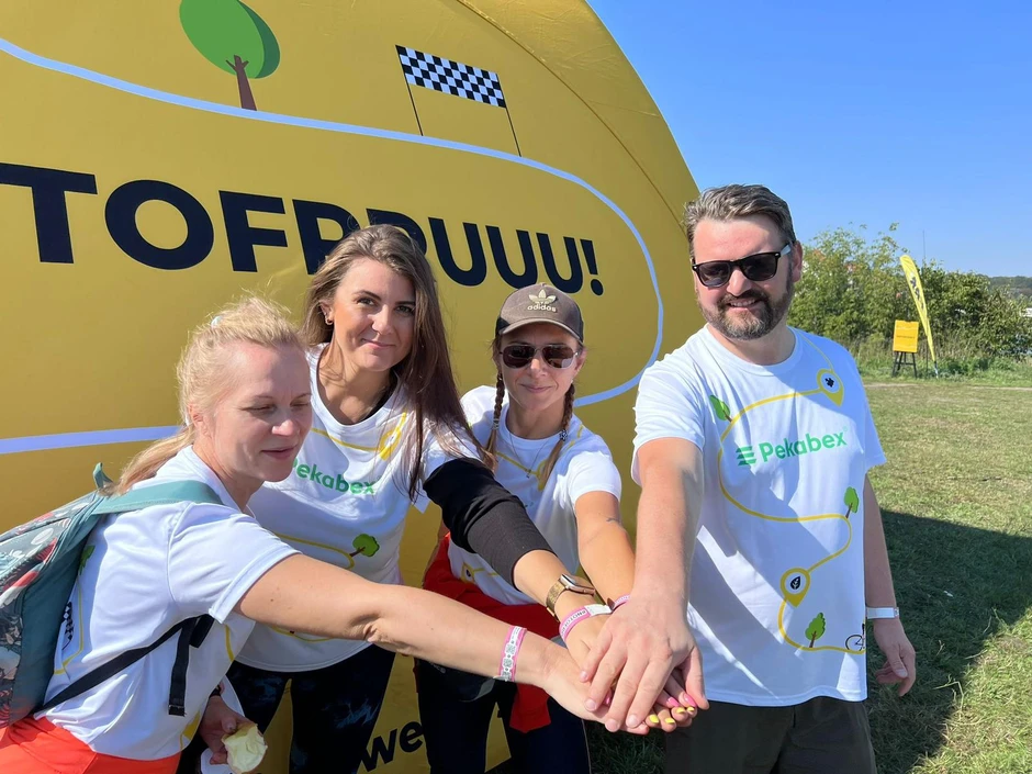 Cztery osoby w koszulkach Pekabex stawiają razem dłonie na tle żółtej ściany z napisem TOFRRUU